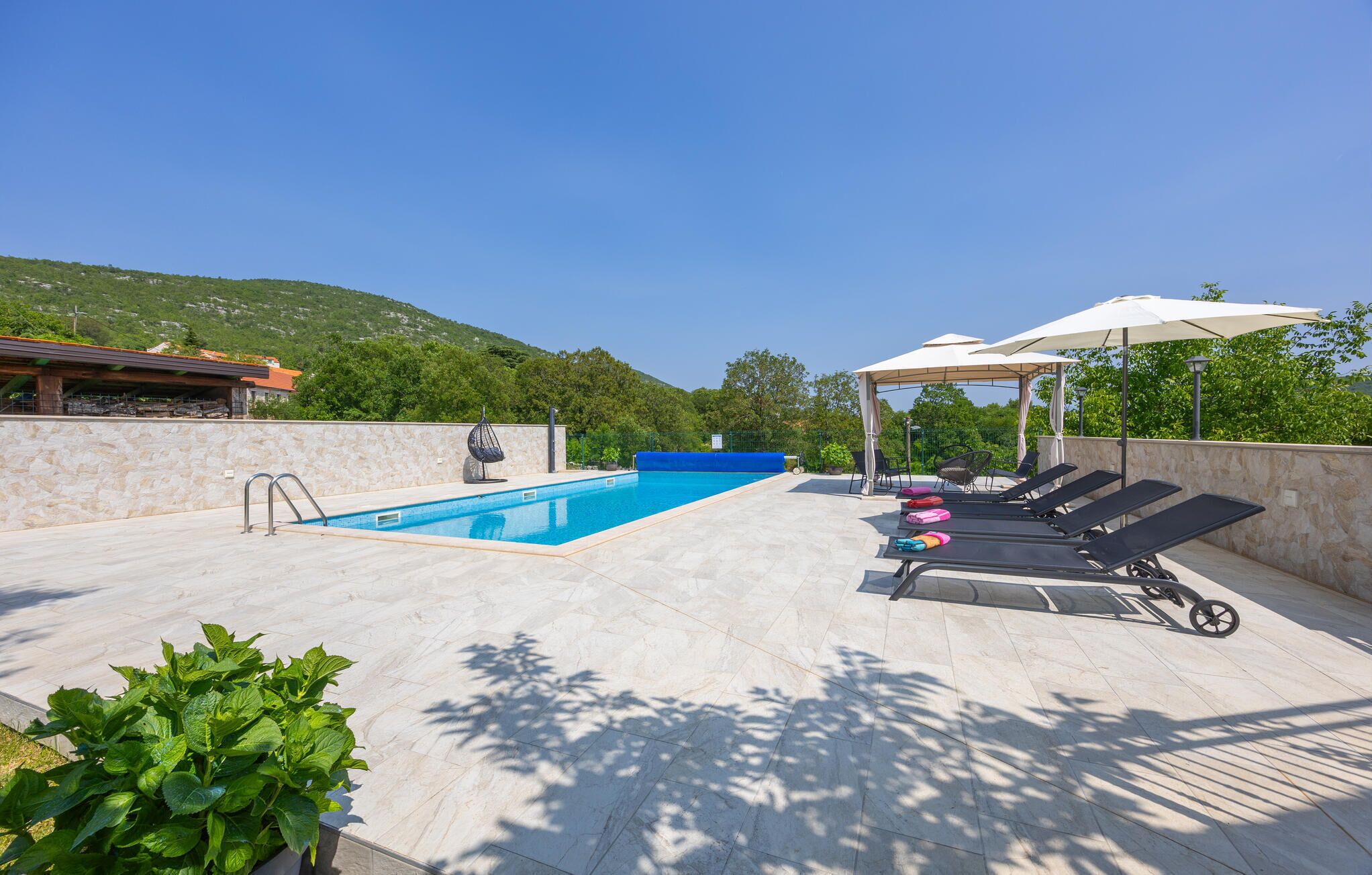 Piscina com espreguiçadeiras e guarda-sóis, rodeada por muros de pedra e vegetação.