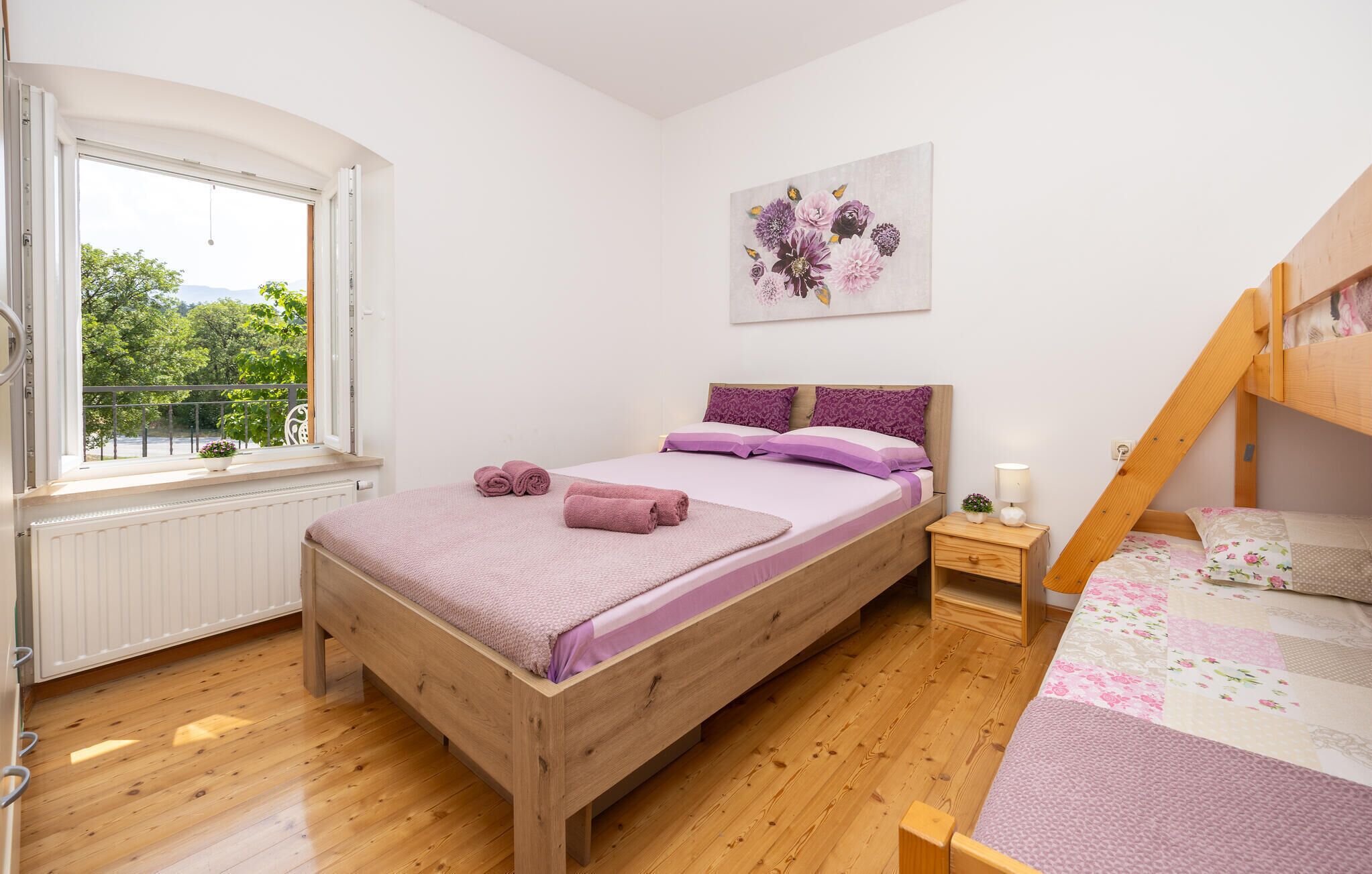A bedroom with a wooden bed, a window with a view of greenery, a small bedside table with a lamp, and a bunk bed in the corner.