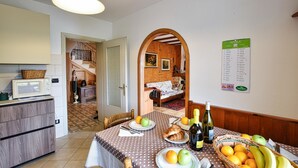 Een eethoek met een tafel gedekt voor twee personen, compleet met fruit, brood en wijn. Op de achtergrond is een keuken zichtbaar met een magnetron en een mand.