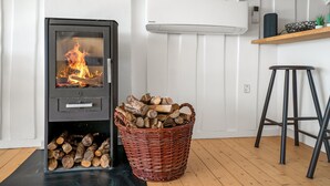 A modern kitchen with a wood-burning stove, a wicker basket of firewood, a bar stool, and a white wall with a built-in air conditioning unit.