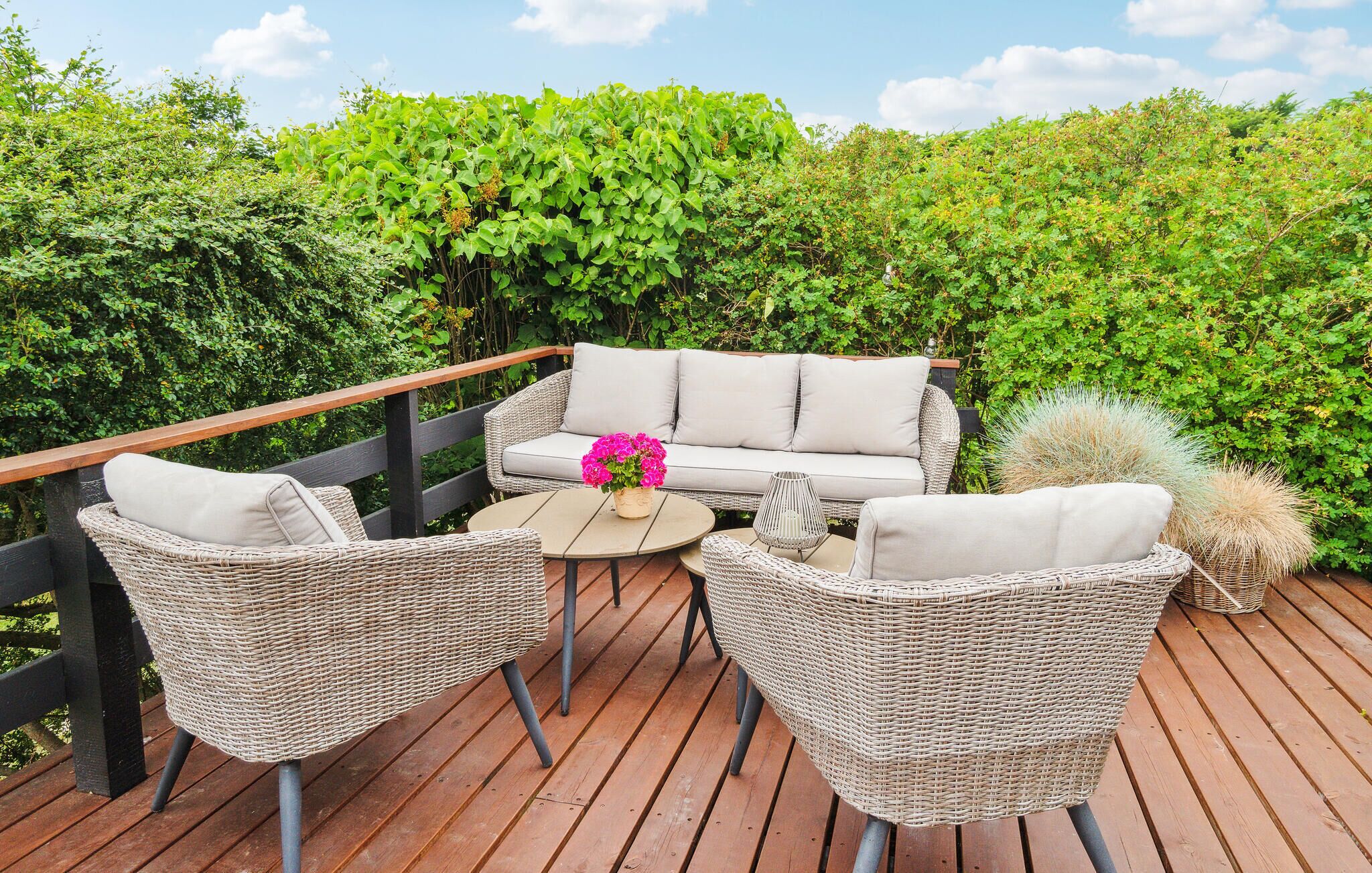 Una terraza al aire libre con mobiliario de mimbre, incluyendo dos sillones y un sofá, una mesa redonda y una maceta con flores rosadas.