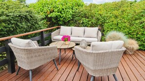 An outdoor deck with wicker furniture, including two chairs and a sofa, a round table, and a potted plant with pink flowers.