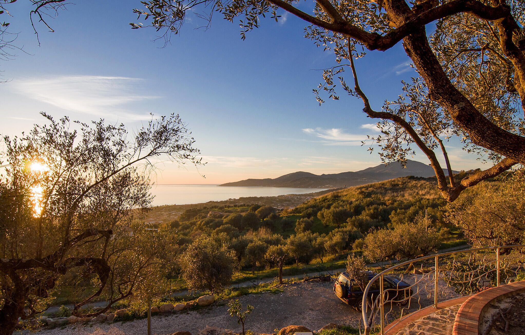 Vista panoràmica d'un paisatge costaner amb cel serè, oliveres i una cadena de muntanyes a la distància.