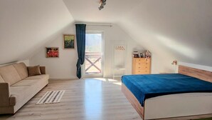 Un dormitori amb sòl de fusta, un llit amb mantell blau, un sofà, un taulell i una finestra amb vista a l'exterior.