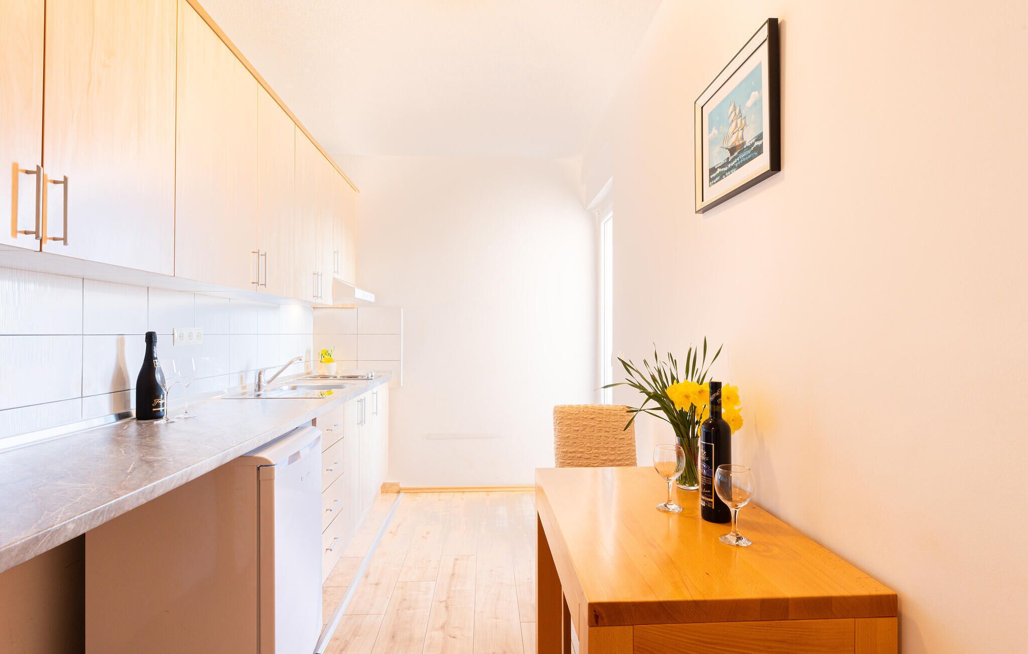 A modern kitchen with white cabinets, a wooden countertop, and a dining area with a wooden table and chairs. There is a framed picture on the wall and a vase with flowers on the table.