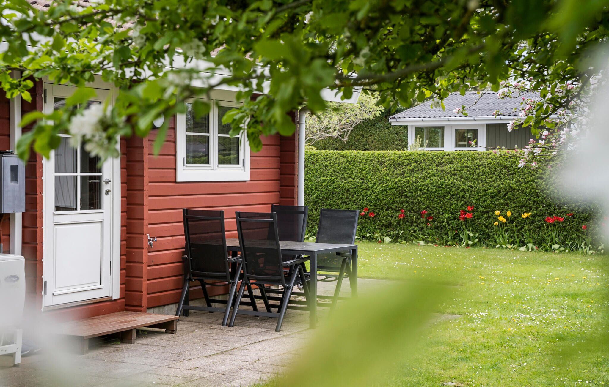Een rood huis met een witte deur en ramen, een klein terras met een tafel en stoelen, en een tuin met diverse bloemen en een haag.