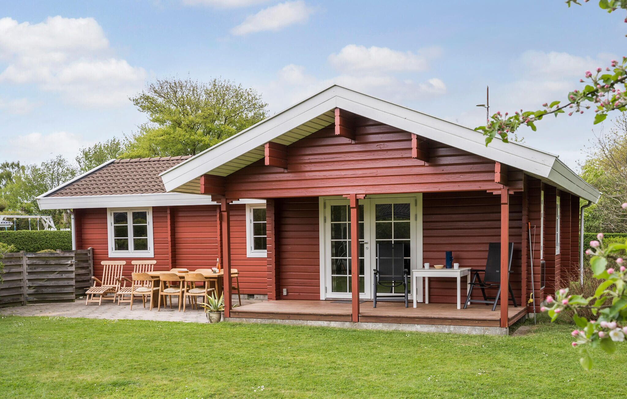 A red wooden house with a porch, a dining area with chairs and a table, and a small garden.