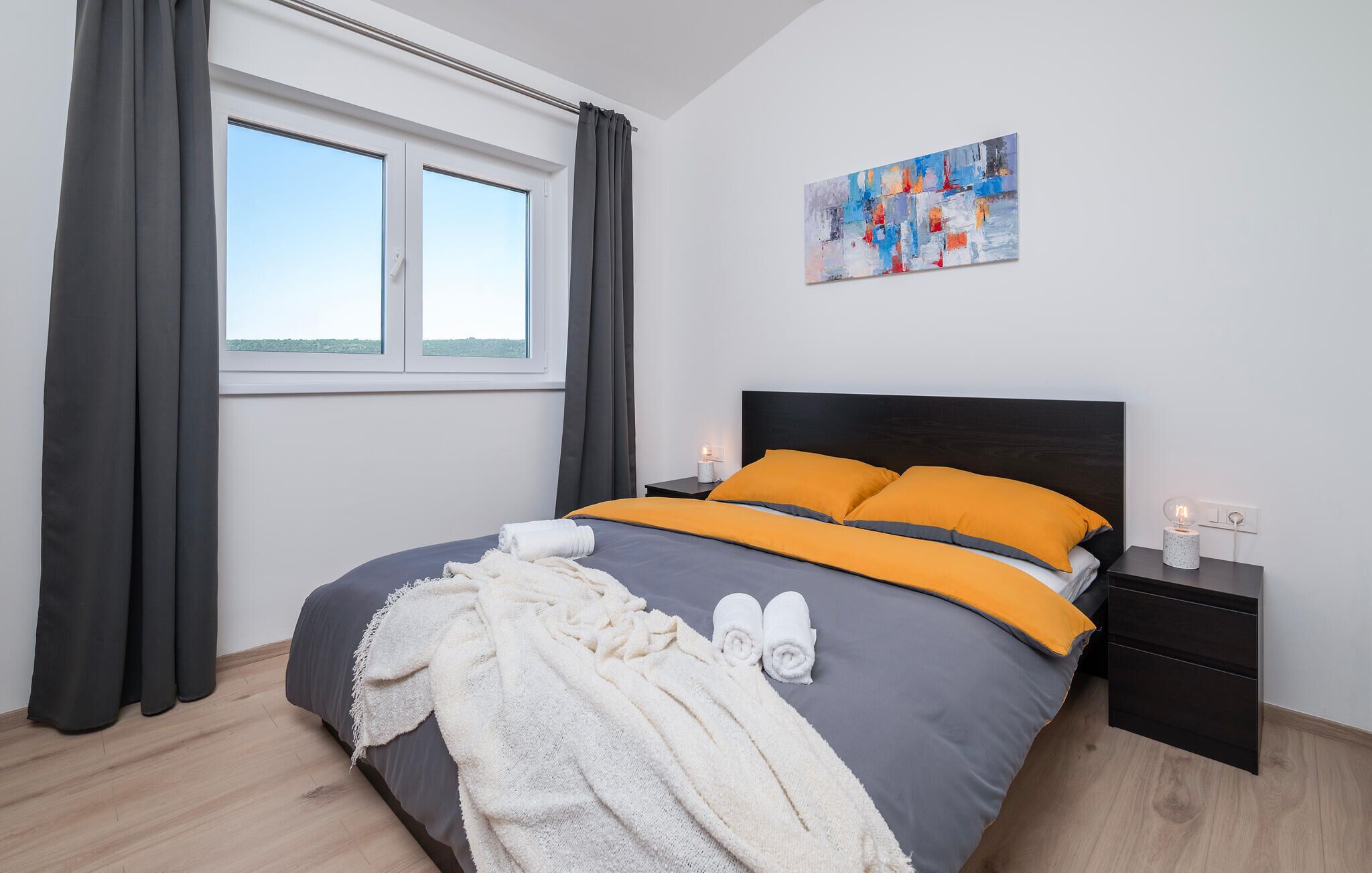 A modern bedroom with a large bed, grey and orange bedding, a white throw blanket, and a painting on the wall.