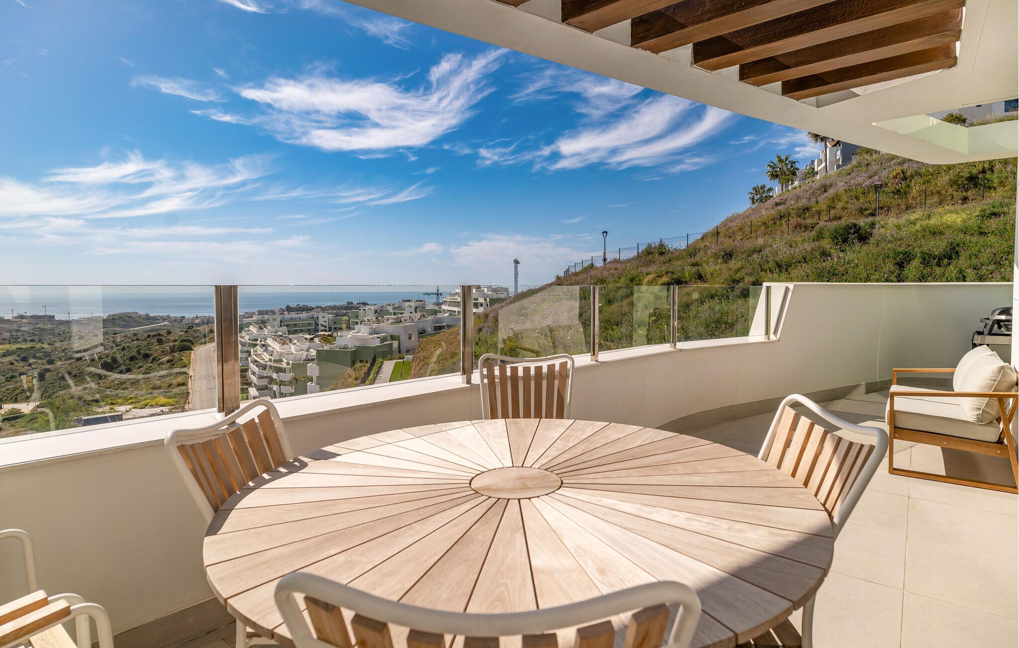A round wooden table with chairs on a balcony overlooking a coastal cityscape.