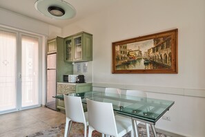 A dining area with a glass table, white chairs, a green kitchen, and a painting of a canal scene.