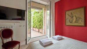 A hotel room with a bed, a chair, a television, a painting, and a view of greenery through an open door.