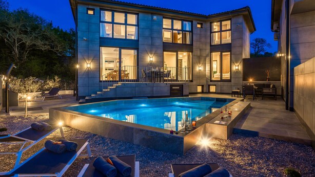 A modern two-story house with a swimming pool, outdoor seating, and a well-lit interior.