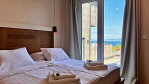 A hotel room with a large bed, two pillows, a nightstand with towels, and a view of the sea through a large window.