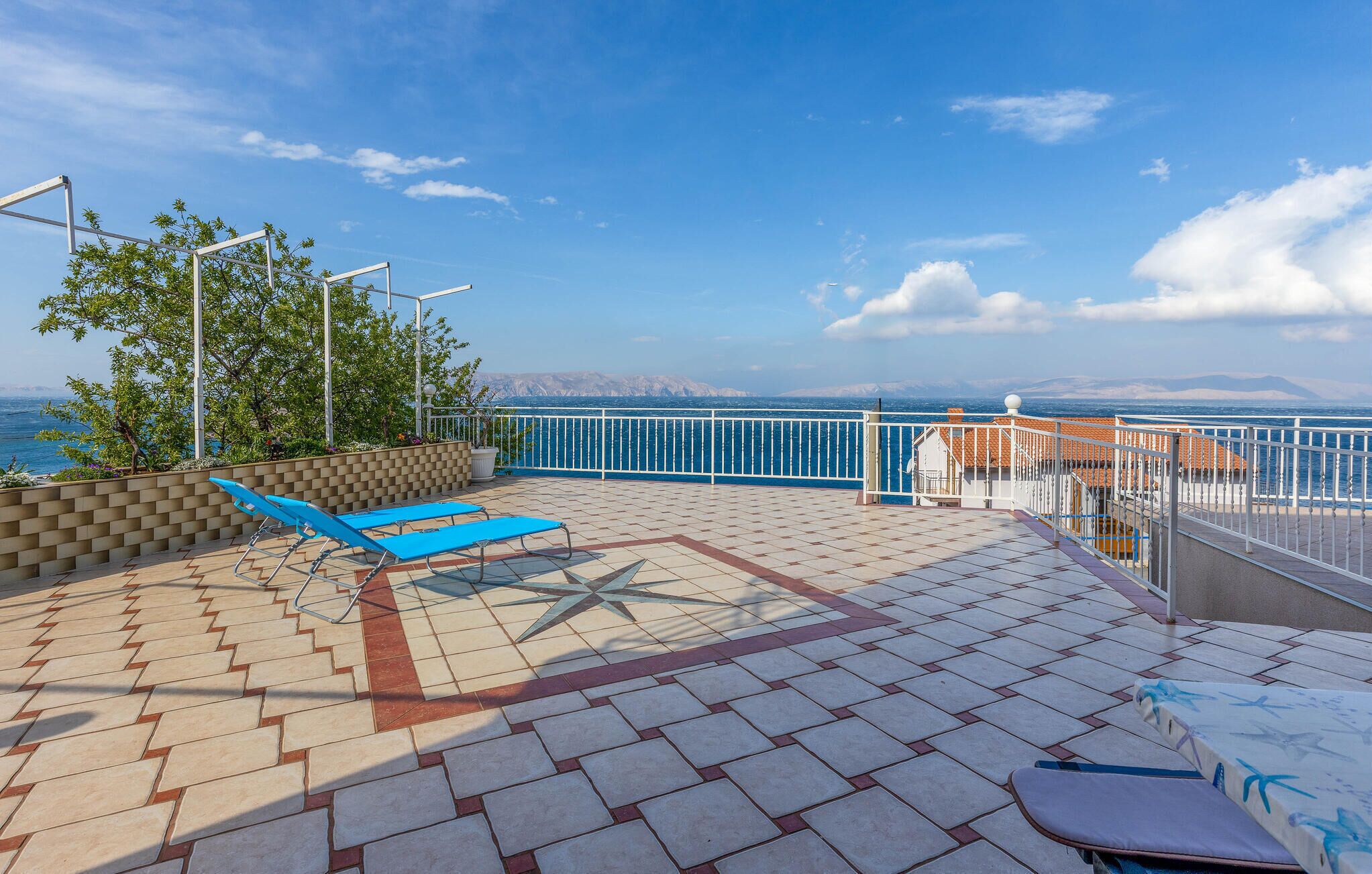Una terraza iluminada por el sol con suelo de baldosas, dos sillones azules, una barandilla blanca y vistas al mar y montañas a lo lejos.