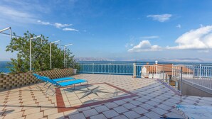 Eine sonnendurchflutete Terrassenanlage mit Fliesenboden, zwei blaue Lounge-Sesseln, einem weißen Geländer und Blick auf das Meer und ferne Berge.
