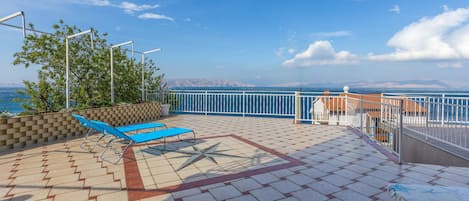 Una terraza iluminada por el sol con suelo de baldosas, dos sillones azules, una barandilla blanca y vistas al mar y montañas a lo lejos.