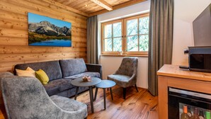Een knus interieur van een chalet met houten plafond, een bank, twee fauteuils, een kleine tafel, een raam met gordijnen, en een keuken met een magnetron.