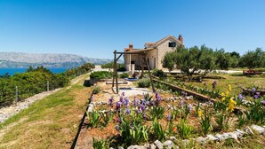 A stone house with a terrace, surrounded by a garden with purple and yellow flowers, and a view of the sea and mountains in the background.