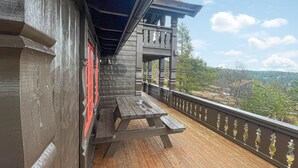 A wooden balcony with a wooden bench, a railing with decorative elements, and a view of a forested landscape.