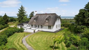 A large white house with a thatched roof, surrounded by lush greenery and a view of the sea. - Stunning home in Fjerritslev with sauna (Fjerritslev)
