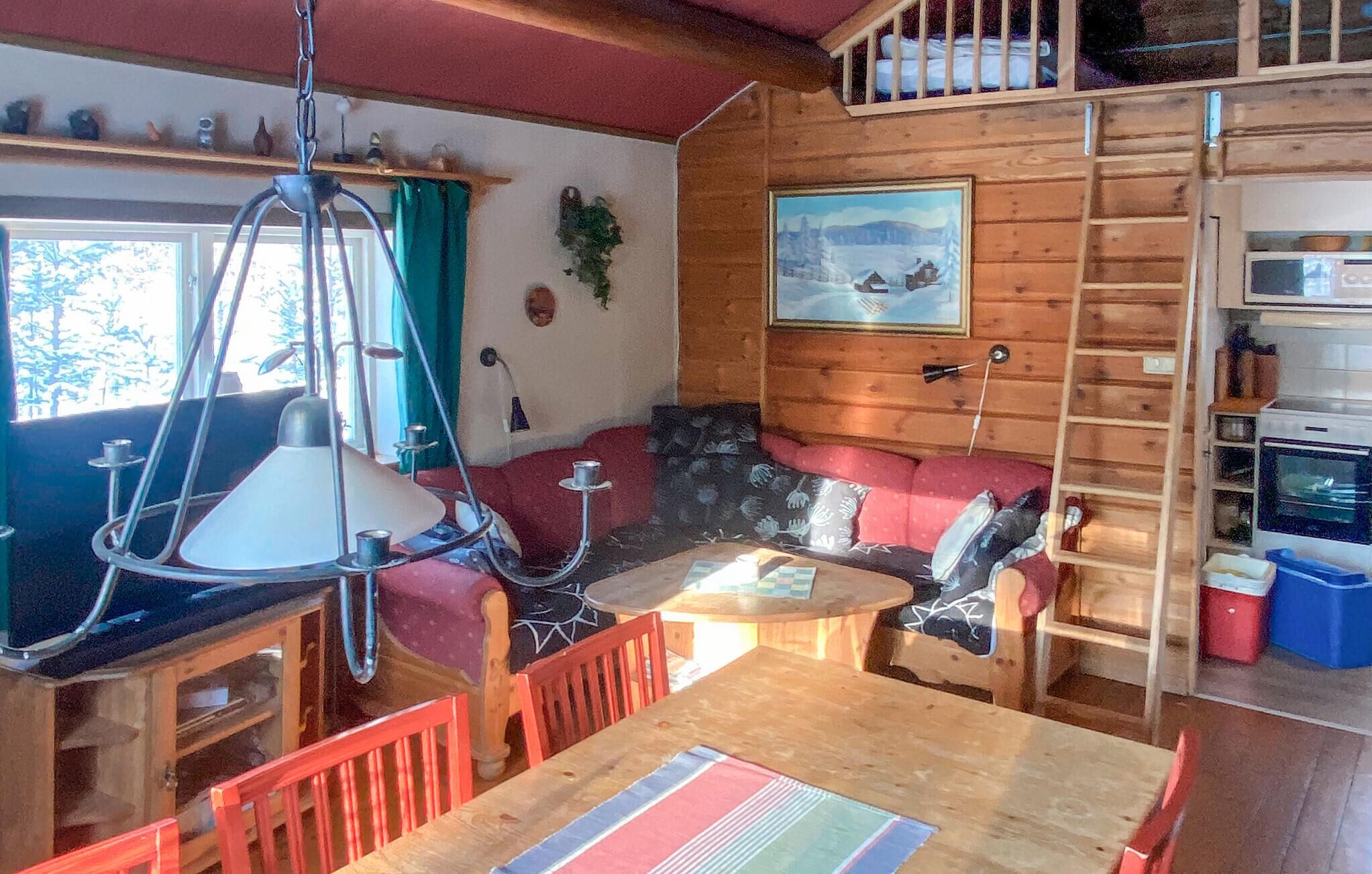 L’intérieur chaleureux d’un chalet avec une table à manger en bois, des chaises rouges, un luminaire suspendu, un canapé avec des coussins à motifs, un tableau représentant un paysage enneigé et un coin cuisine équipé d’un micro-ondes et de quelques étagères.