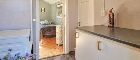 Een compacte keuken met een stenen aanrechtblad, een wit kastje en een deur naar een slaapkamer met een bed en nachtkastje.