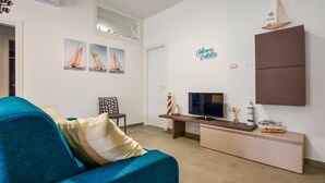 Uma sala de estar com um sofá azul-esverdeado, uma TV sobre um suporte de madeira e uma parede com pinturas de veleiros e uma placa.