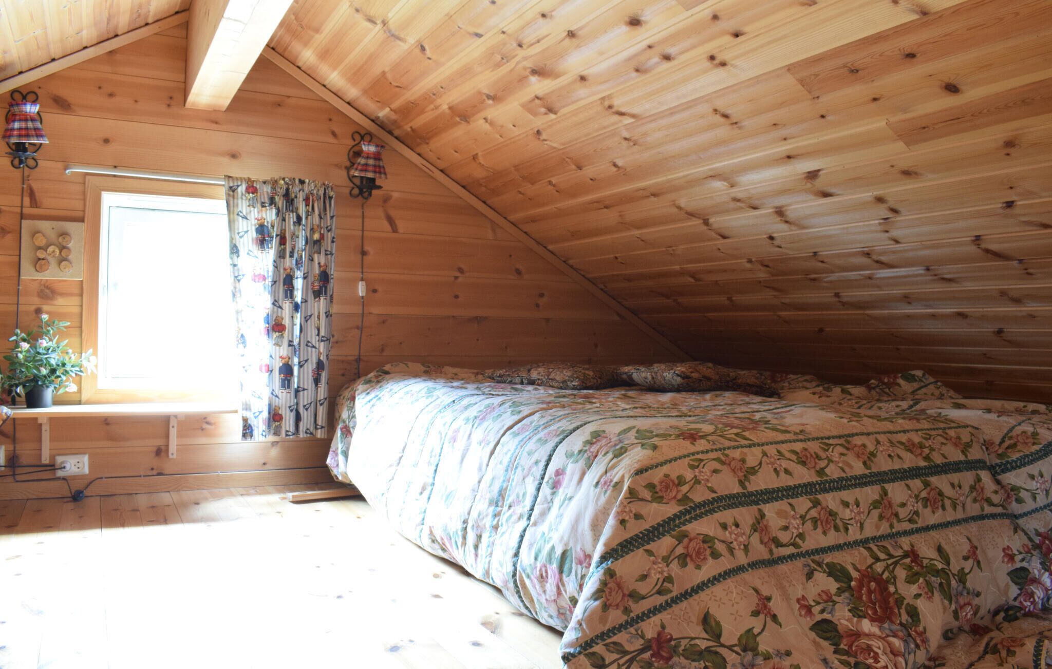 A cozy attic bedroom with a wooden sloped ceiling, a bed with floral bedding, a window with curtains, and a small table with a plant.