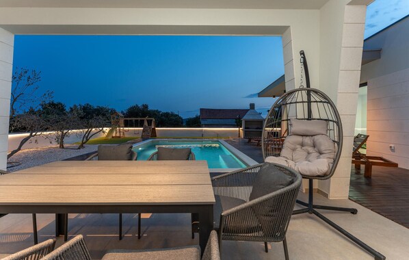 A poolside area with a wooden table, wicker chairs, a hanging chair, and a children's playset.