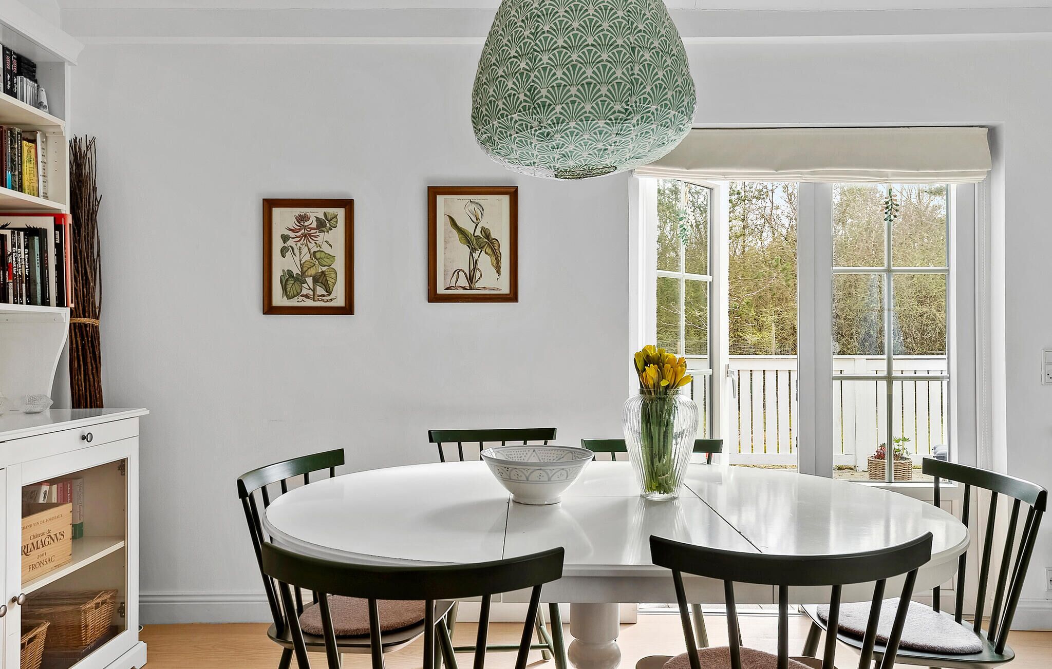 A dining area with a round table, chairs, a vase with flowers, and framed botanical illustrations on the wall.