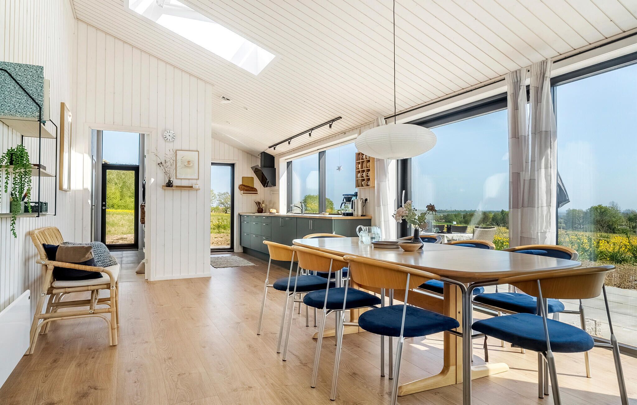 A spacious dining area with a large wooden table and blue upholstered chairs, a kitchen with a view, and a white interior with wooden beams.