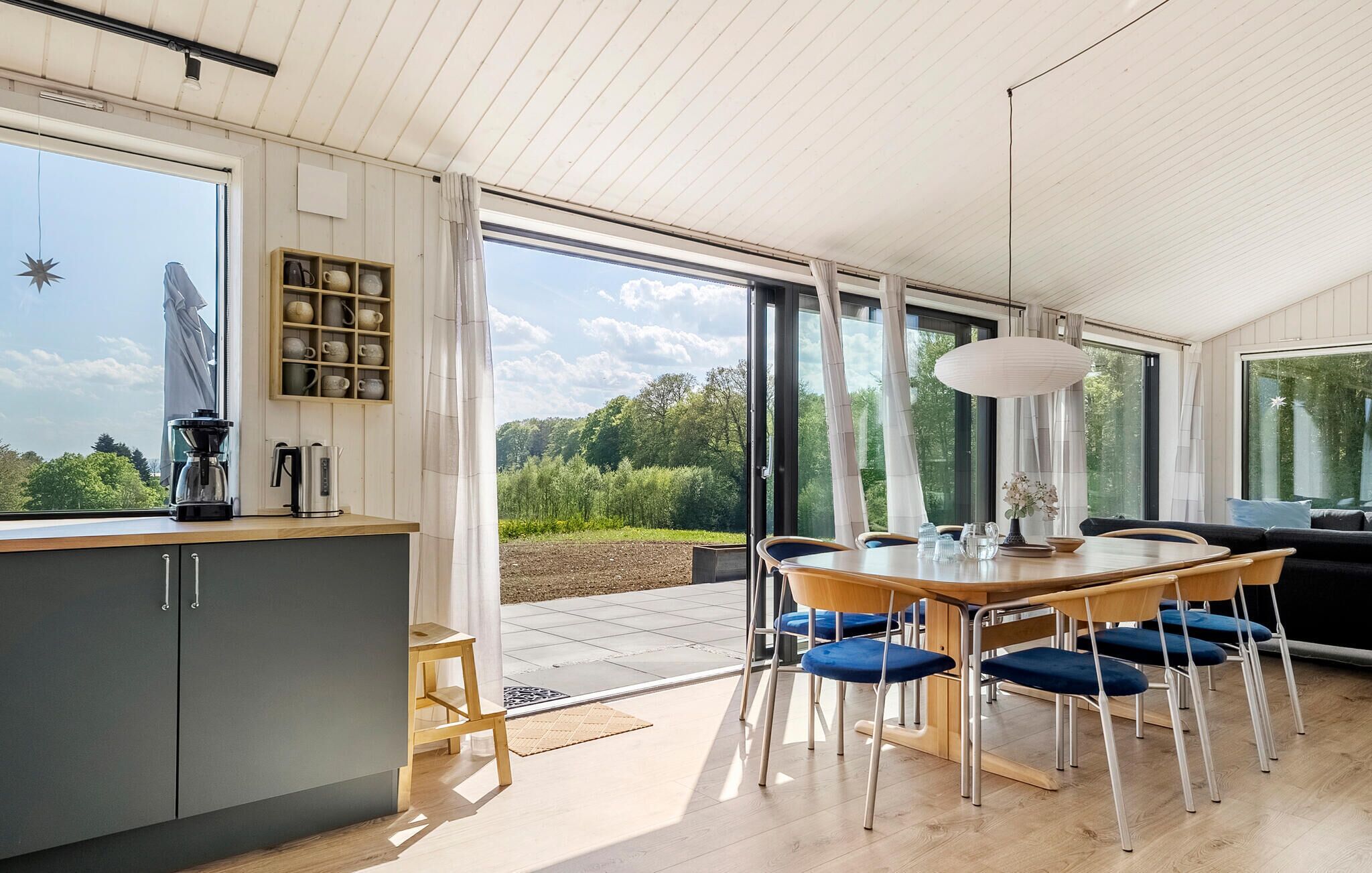 A modern kitchen with a wooden countertop, a coffee maker, and a kettle. There is a dining area with a wooden table and blue chairs. The room has large windows with a view of greenery outside.