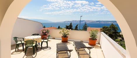 Een balkon met een tafel en stoelen, bloempotten en uitzicht op zee en bergen.