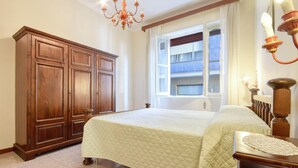 A bedroom with a large bed, wooden wardrobe, and a window with a view of buildings.
