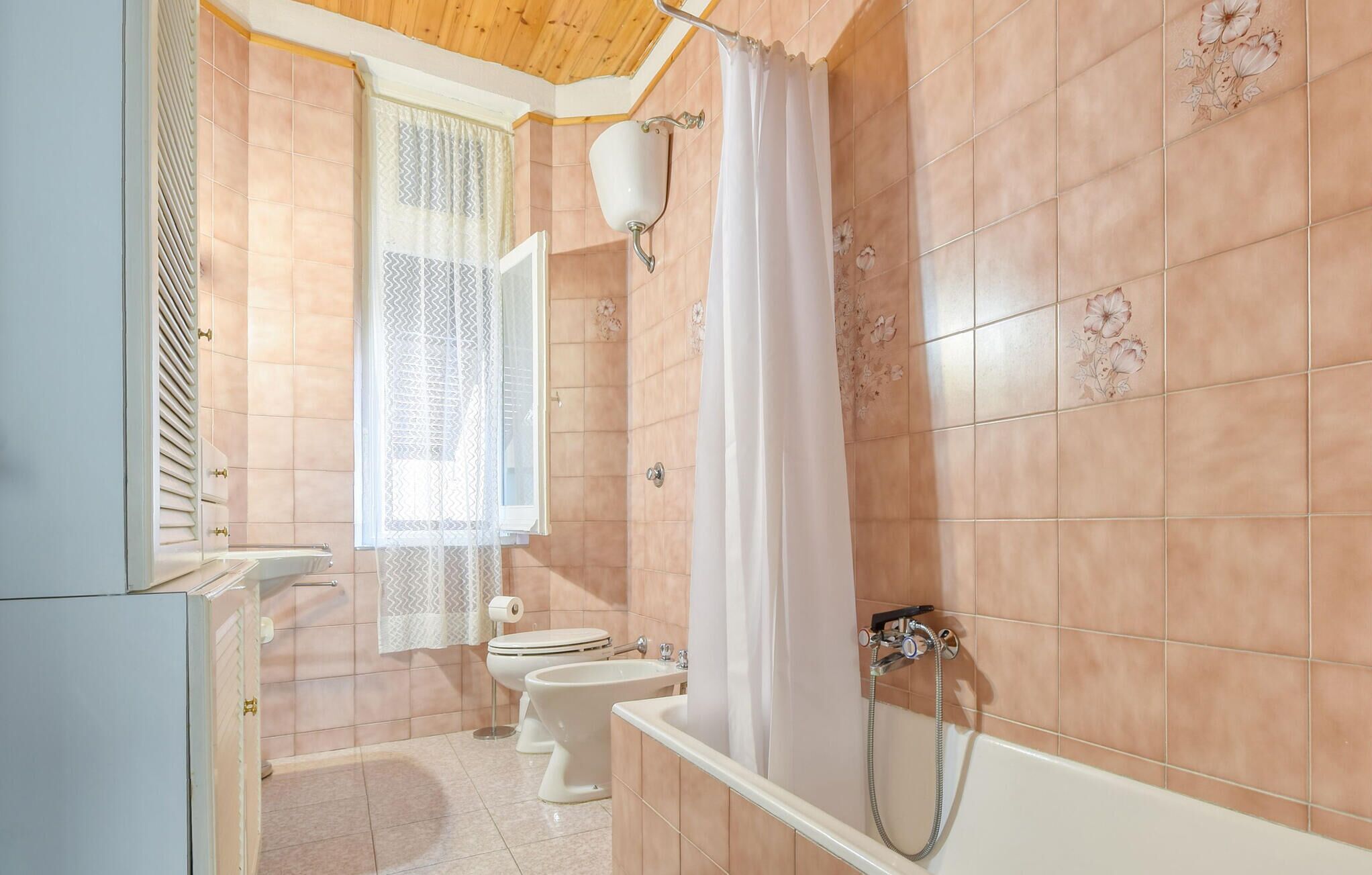 Une salle de bain avec une baignoire, deux toilettes, une fenêtre avec un rideau à motifs et un luminaire fixé au mur.