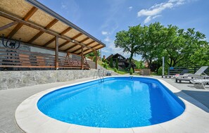 A swimming pool with a stone wall and wooden deck, surrounded by trees and a building with a wooden roof. - Beautiful home in Radoboj (Radoboj)