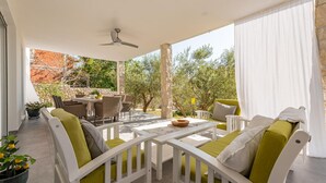 Un espacio de estar al aire libre techado con mobiliario blanco y una mesa, rodeado de follaje y un muro de piedra.