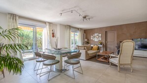 A modern living room with a glass dining table, two sofas, a TV, and a large window with curtains.