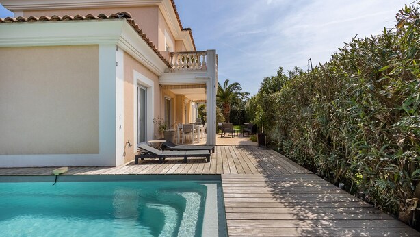 A wooden deck leading to a swimming pool with lounge chairs, a patio area, and dense foliage on one side.