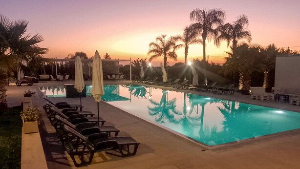Ein Schwimmbecken mit Liegestühlen und Sonnenschirmen, umgeben von Palmen und Blick auf den Sonnenuntergang.