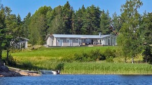 Suuri, yhden kerroksen talo, jossa on katettu kuisti, ja joka sijaitsee ruohikkomäellä ja josta avautuu näkymä vesistöön.