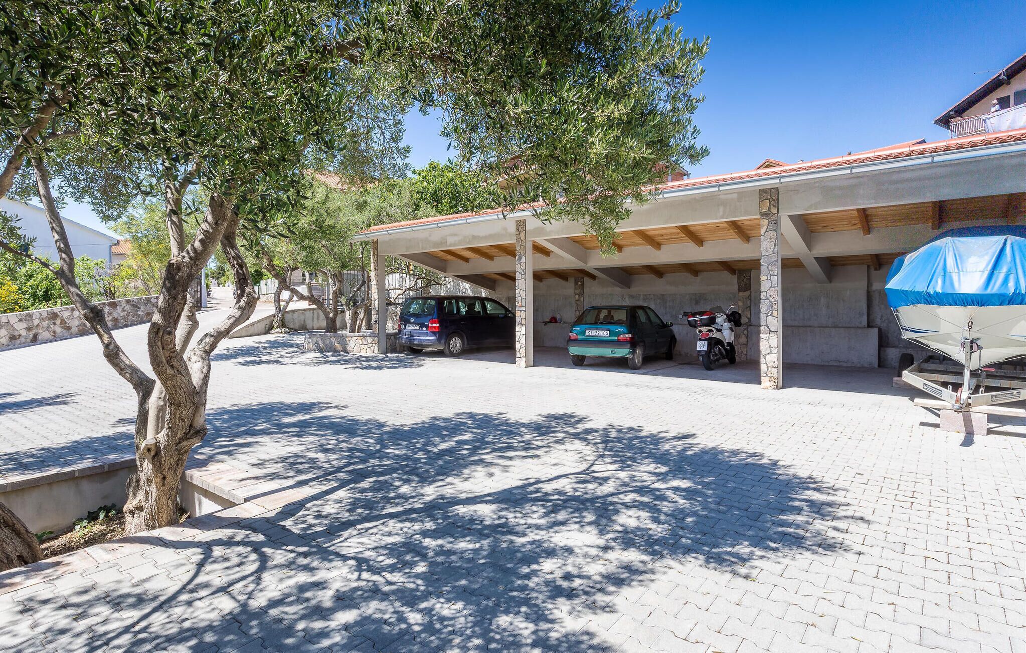 A covered parking area with two cars parked under a roof, a tree in the foreground, and a building with a tiled roof in the background.