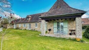 A stone house with a steeply pitched roof, a small porch with outdoor seating, and a well-maintained lawn.