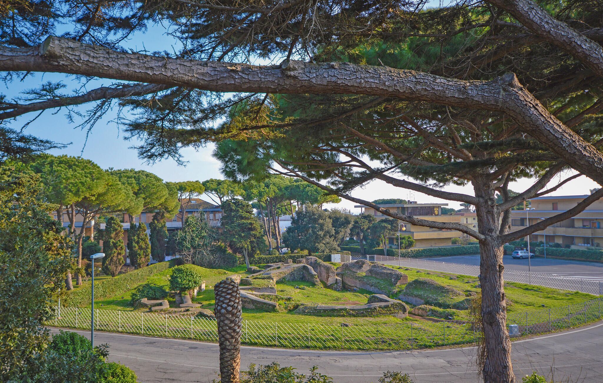 Un parque con ruinas antiguas, rodeado de árboles y edificios modernos al fondo.