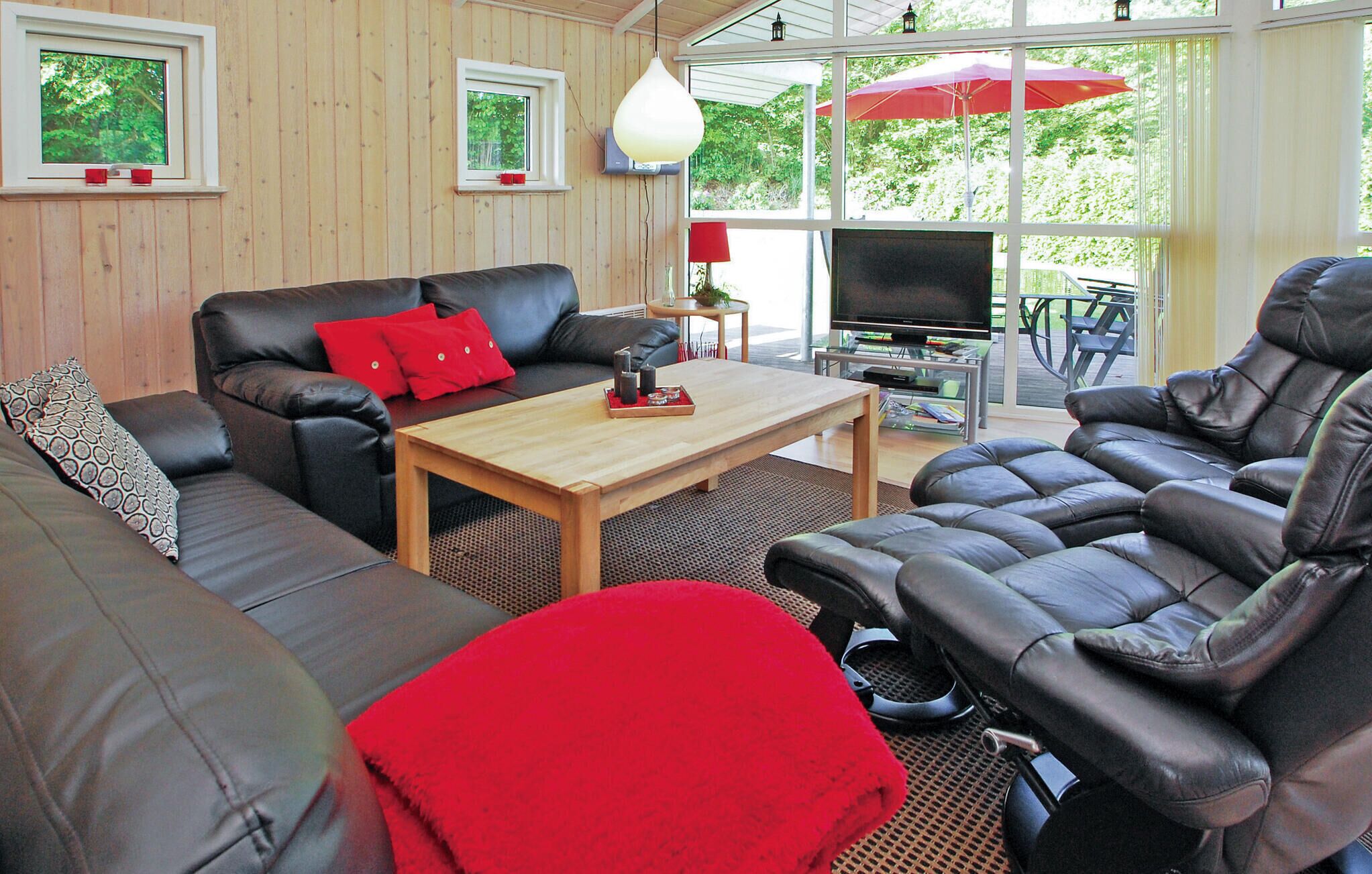 Ein gemütliches Wohnzimmer mit einer Couch, einem Couchtisch und einem Fernseher. Es gibt Fenster mit Vorhängen und einen Blick ins Grüne.