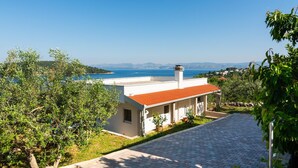 A white building with a red roof, surrounded by greenery and overlooking a coastal view.