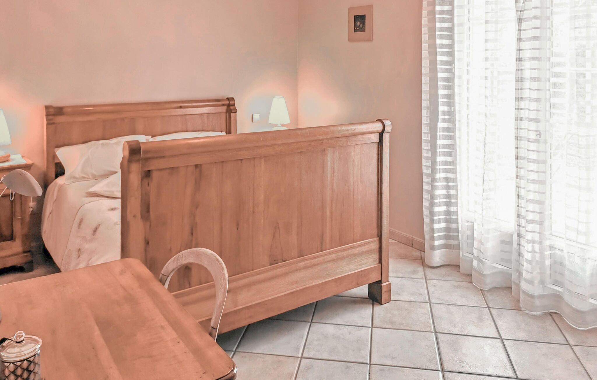 A bedroom with a wooden bed, a bedside table, a lamp, and a window with sheer curtains.