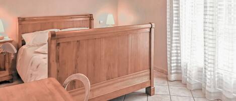 A bedroom with a wooden bed, a bedside table, a lamp, and a window with sheer curtains.