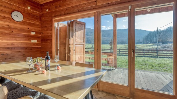 Interior de madera con una mesa de comedor para dos personas, un reloj en la pared y puertas correderas de cristal que dan a un área exterior con césped.