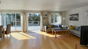 A spacious living room with a wooden floor, a sofa, a coffee table, and a TV. There is a balcony with a railing and a view of trees.